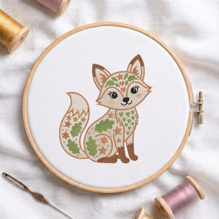 A circular embroidery hoop holding white fabric with a stylized cartoon fox design filled with leaf and star patterns in green, tan, and brown. The hoop is photographed on a white fabric surface with spools of thread and a needle around it.