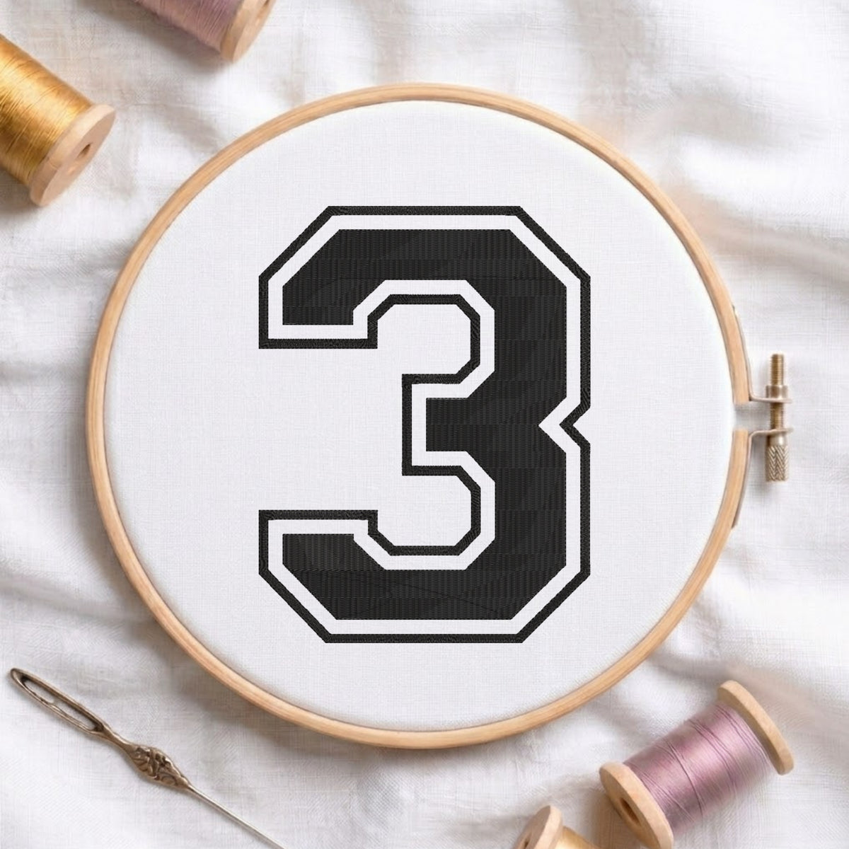 A wooden embroidery hoop holding white fabric with a large black number 3 outlined in white. The hoop is styled on a white fabric surface with spools of thread and a needle nearby.