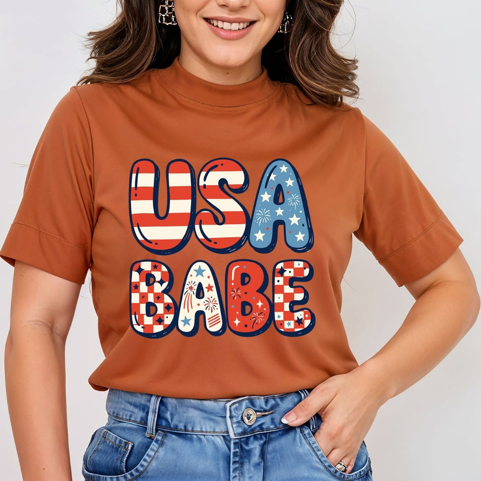 A woman wearing a rust-orange short-sleeve t-shirt with a patriotic graphic that reads 'USA BABE' in red, white, and blue patterned letters, paired with blue jeans.