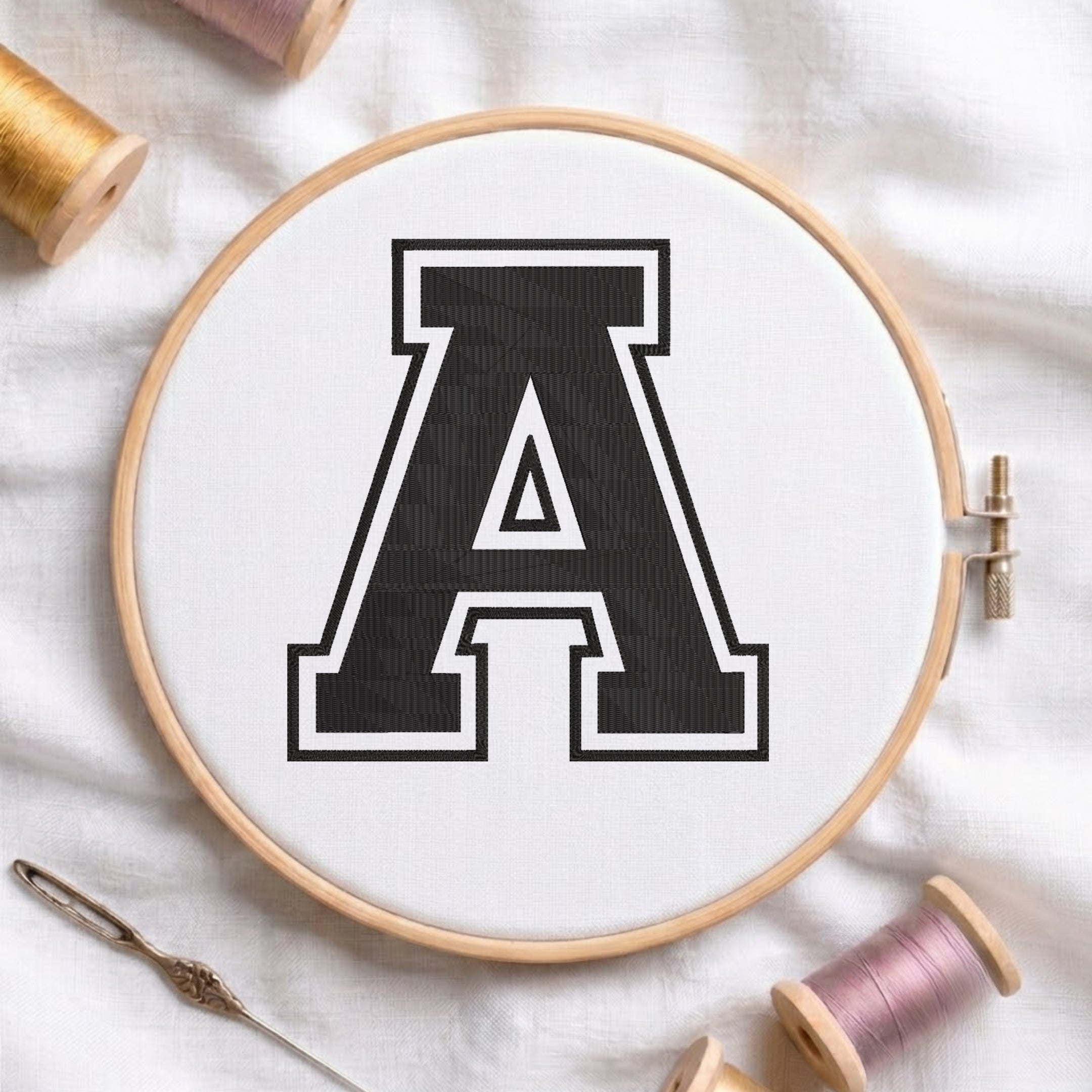 A wooden embroidery hoop holding white fabric with a large black letter A outlined in white, styled on a white fabric background with spools of thread and a needle.