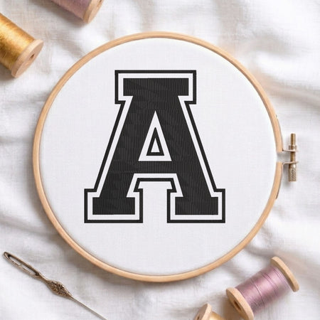 A wooden embroidery hoop holding white fabric with a large black letter A outlined in white, styled on a white fabric background with spools of thread and a needle.