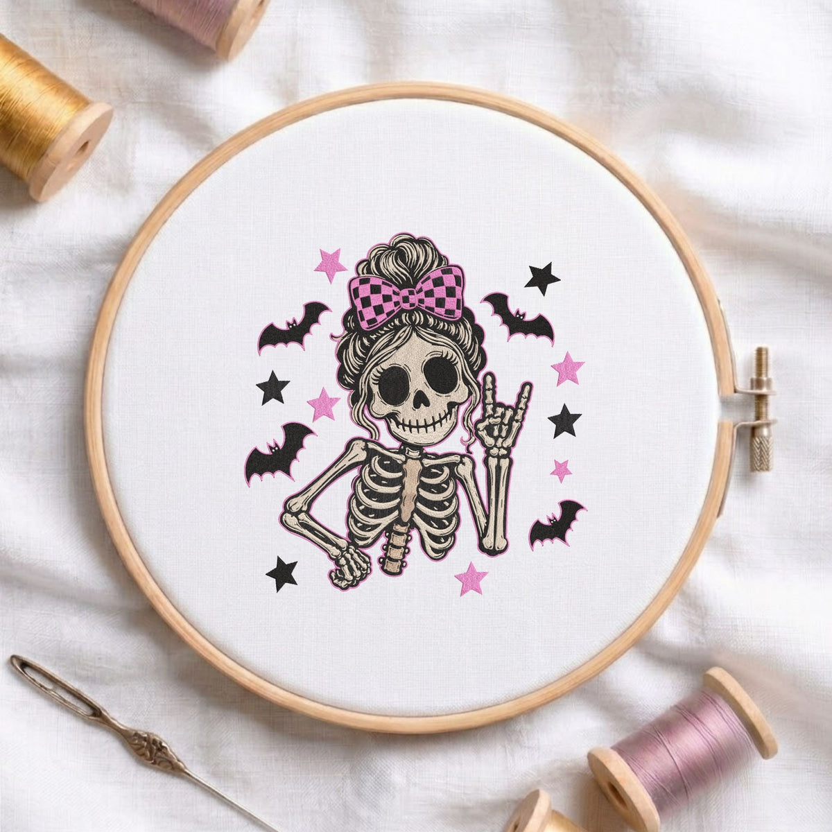 Cute skeleton wearing a messy bun with a pink checkered bow and making a rock sign with bats and stars machine embroidery design shown in a wooden embroidery hoop on white fabric with thread spools and a needle