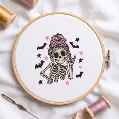 Cute skeleton wearing a messy bun with a pink checkered bow and making a rock sign with bats and stars machine embroidery design shown in a wooden embroidery hoop on white fabric with thread spools and a needle