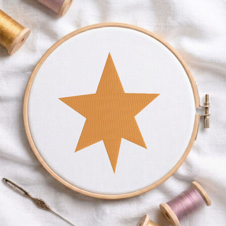 Seven-pointed gold star machine embroidery design displayed inside a round wooden hoop on white linen fabric, accompanied by wooden thread spools and a needle.