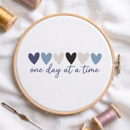 A round embroidery hoop holding white fabric with embroidered six hearts in navy, light blue, beige, black, light gray, and medium blue above the phrase “one day at a time” in navy script. The hoop is styled on a white fabric surface with spools of thread, a needle, and a pin around it.