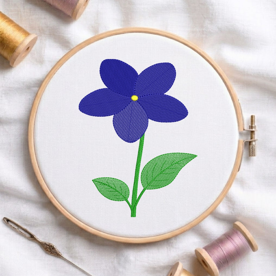 A circular embroidery hoop holding white fabric with a stitched flower design in deep blue, green, and yellow, surrounded by spools of thread and a needle on a white fabric background.