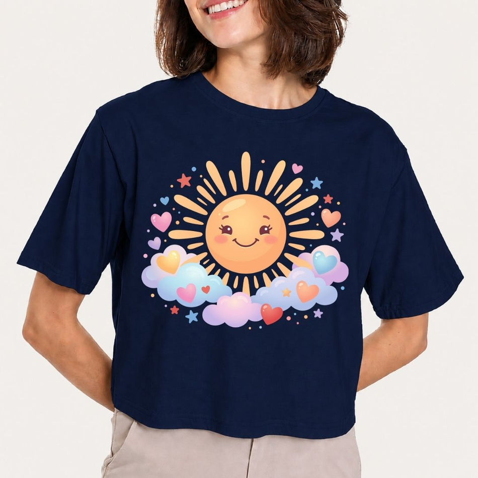 A woman wearing a navy blue short-sleeve t-shirt with a colorful graphic of a smiling sun, pastel clouds, and hearts on the front, paired with beige pants against a light gray background.