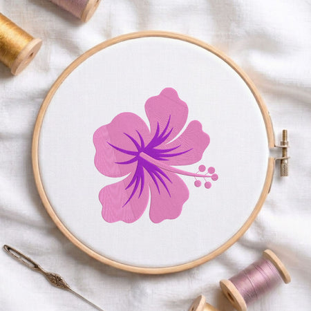 A wooden embroidery hoop holding white fabric with a pink hibiscus flower design, surrounded by spools of thread and a needle on a white fabric background.