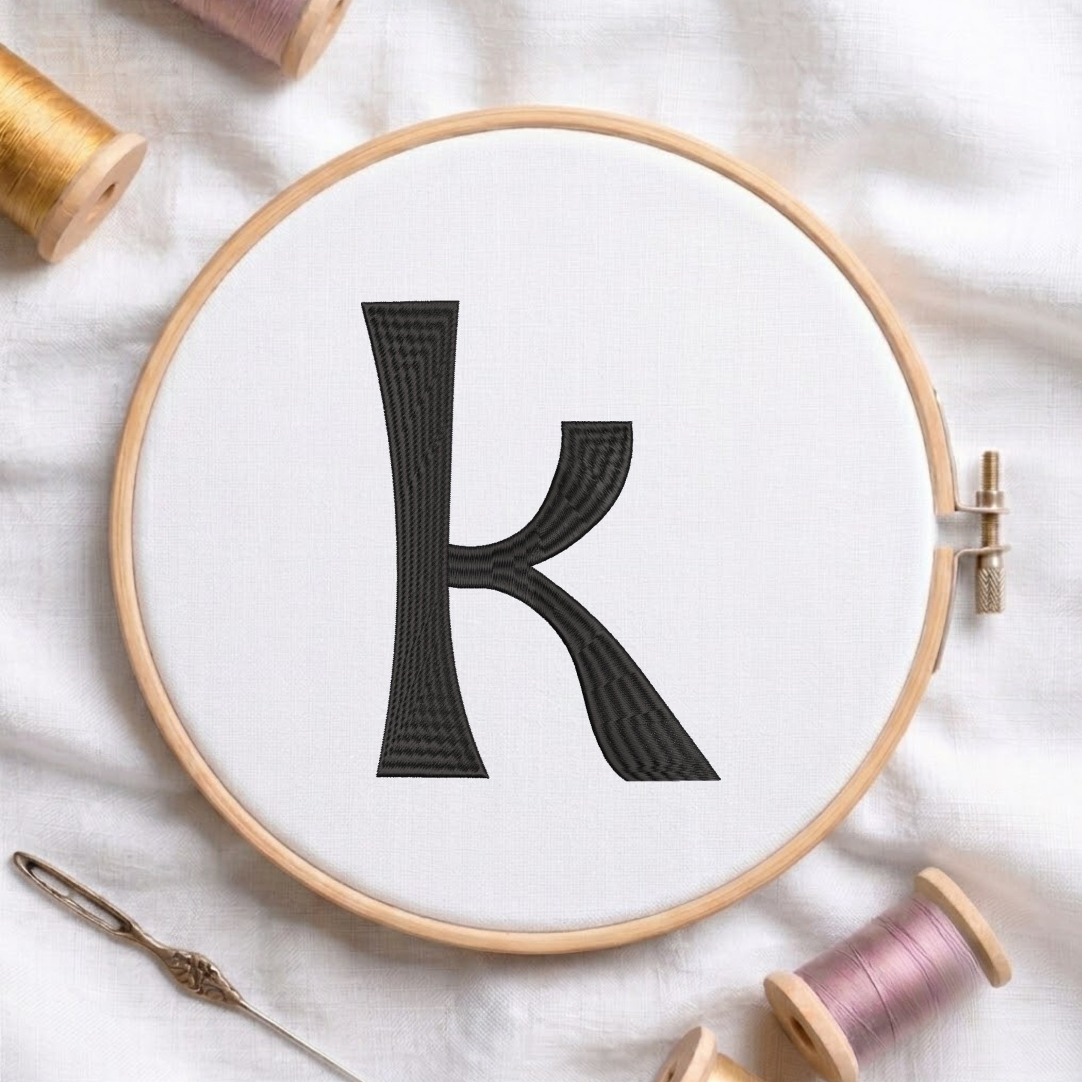 A wooden embroidery hoop holds white fabric with a large black letter 'K' stitched in the center. A metal needle lies nearby on a white fabric surface, with spools of thread partially visible around the edges.