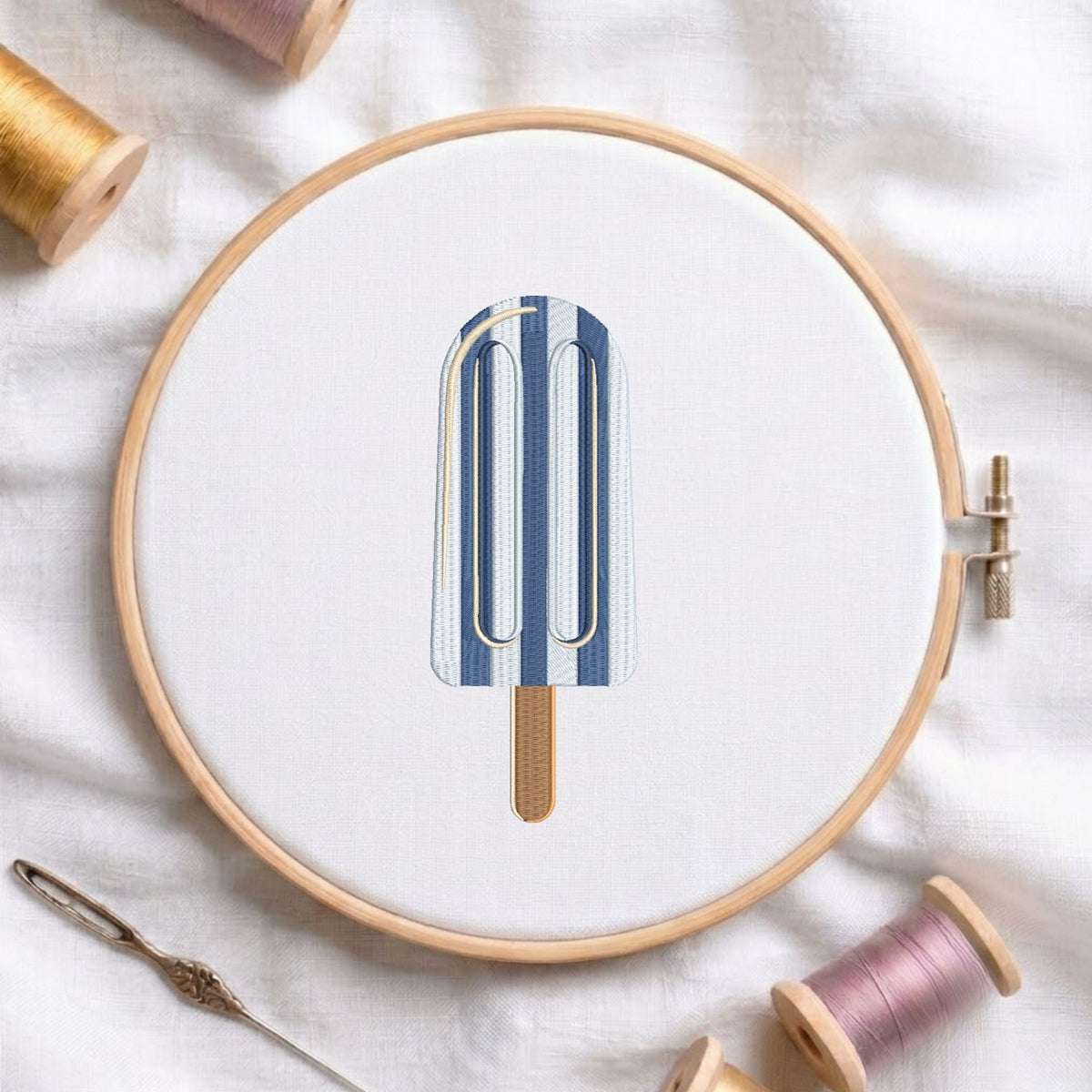 Blue and white striped ice cream popsicle with wooden stick machine embroidery design, displayed inside a round wooden hoop on white fabric with thread spools and a needle.