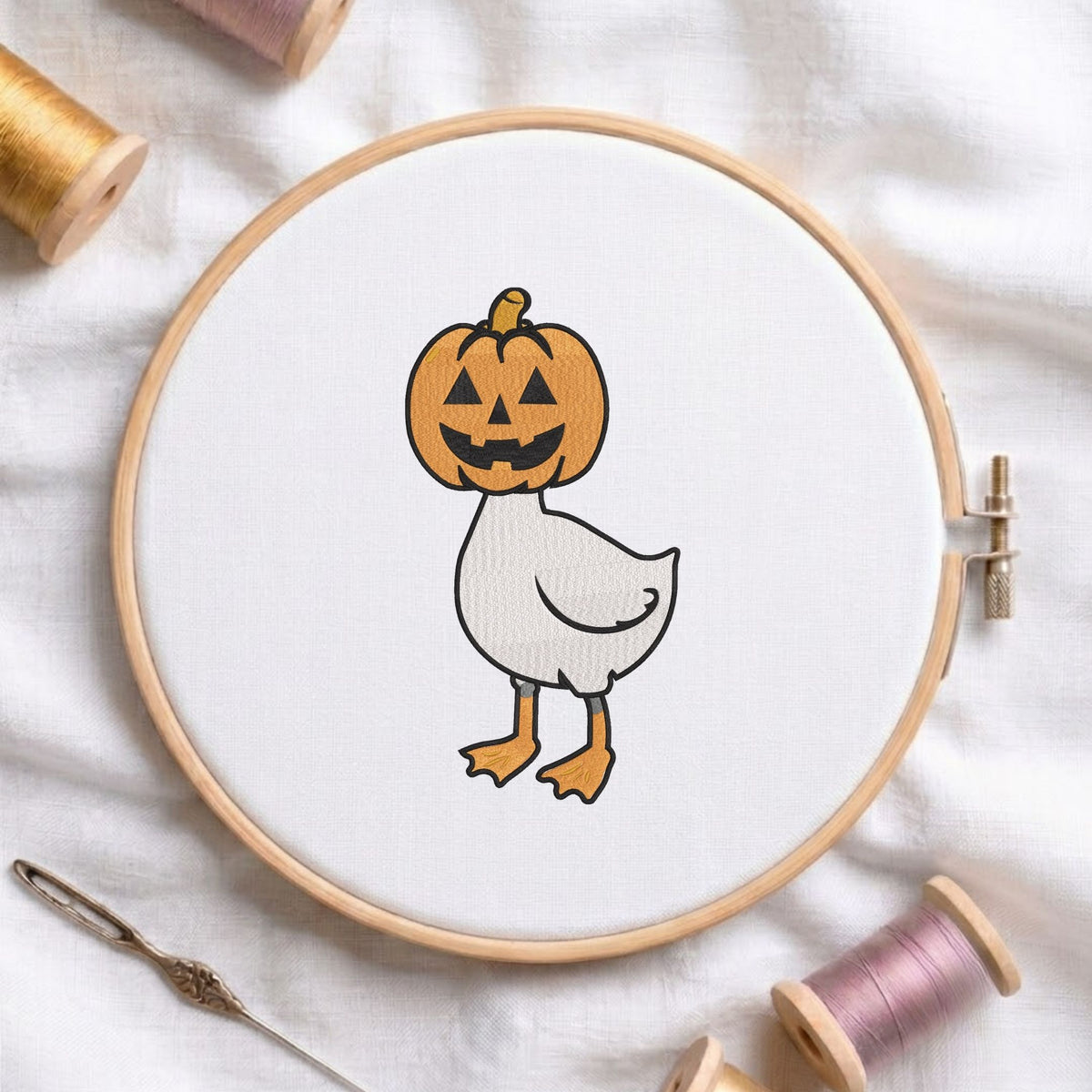 Cute duck wearing a jack-o-lantern pumpkin head for Halloween machine embroidery design shown in a wooden embroidery hoop on white fabric with thread spools and a needle   
