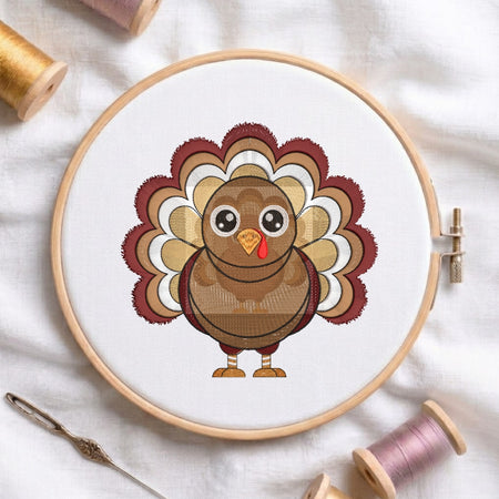 A circular embroidery hoop on white fabric displays a cartoon turkey design with layered brown, tan, cream, and burgundy feathers. Spools of thread and a needle are arranged around it on a white cloth background.