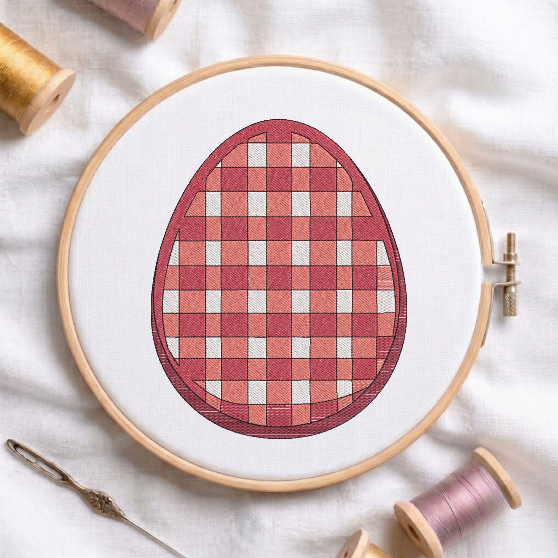 A circular embroidery hoop holding white fabric with a stitched egg design in a red and pink gingham pattern. A wooden needle and spools of thread are arranged around it on a white fabric background.