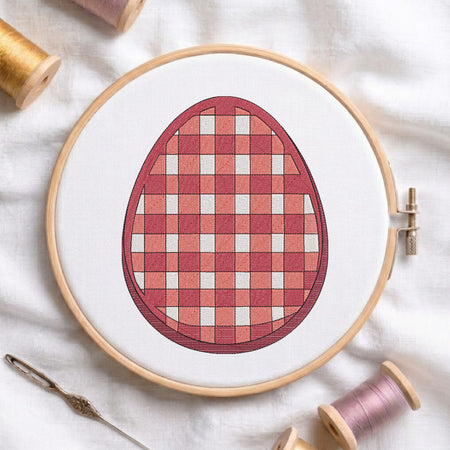 A circular embroidery hoop holding white fabric with a stitched egg design in a red and pink gingham pattern. A wooden needle and spools of thread are arranged around it on a white fabric background.