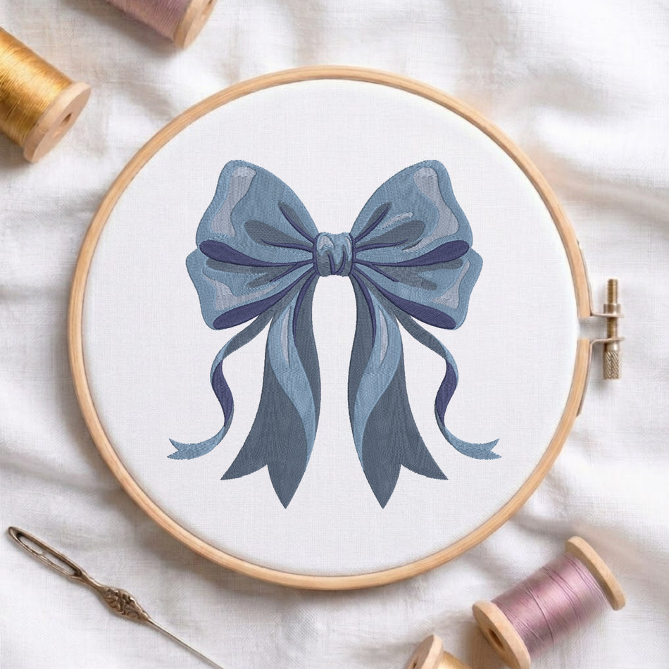Blue bow machine embroidery design displayed inside a round wooden hoop on white linen fabric, accompanied by wooden thread spools and a needle.