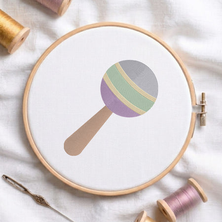 A wooden embroidery hoop holds white fabric with a simple embroidered lollipop design in muted pastel colors. The hoop is styled on a white fabric surface with thread spools and a needle around it.