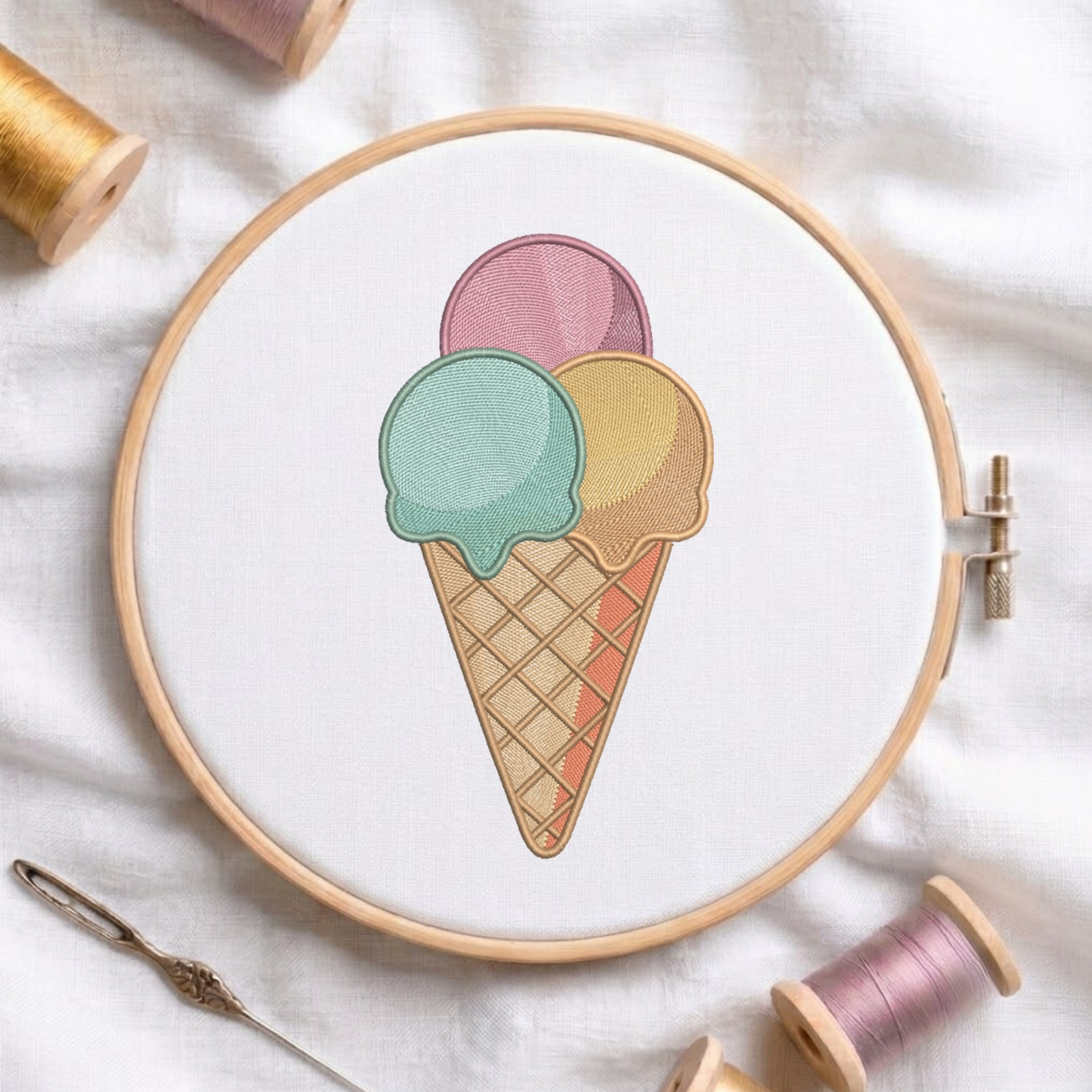 A circular embroidery hoop holding white fabric with a pastel ice cream cone design, surrounded by spools of thread and a needle on a white fabric background.