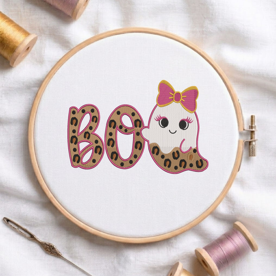 A white fabric stretched in a wooden embroidery hoop, featuring an embroidered design of the text "BOO" in leopard print with pink outlines, alongside a cute ghost wearing a pink bow and leopard print details.