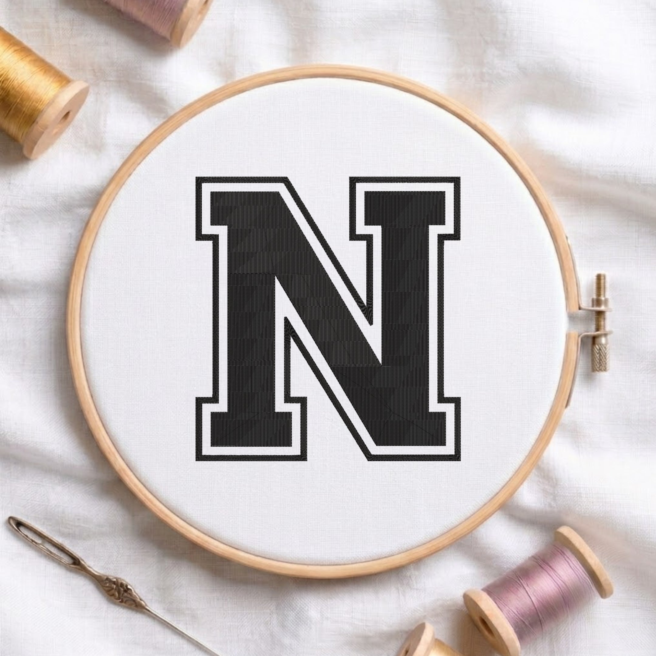 A wooden embroidery hoop holding white fabric with a large black letter N outlined in white, styled on a white fabric background with spools of thread and a needle.