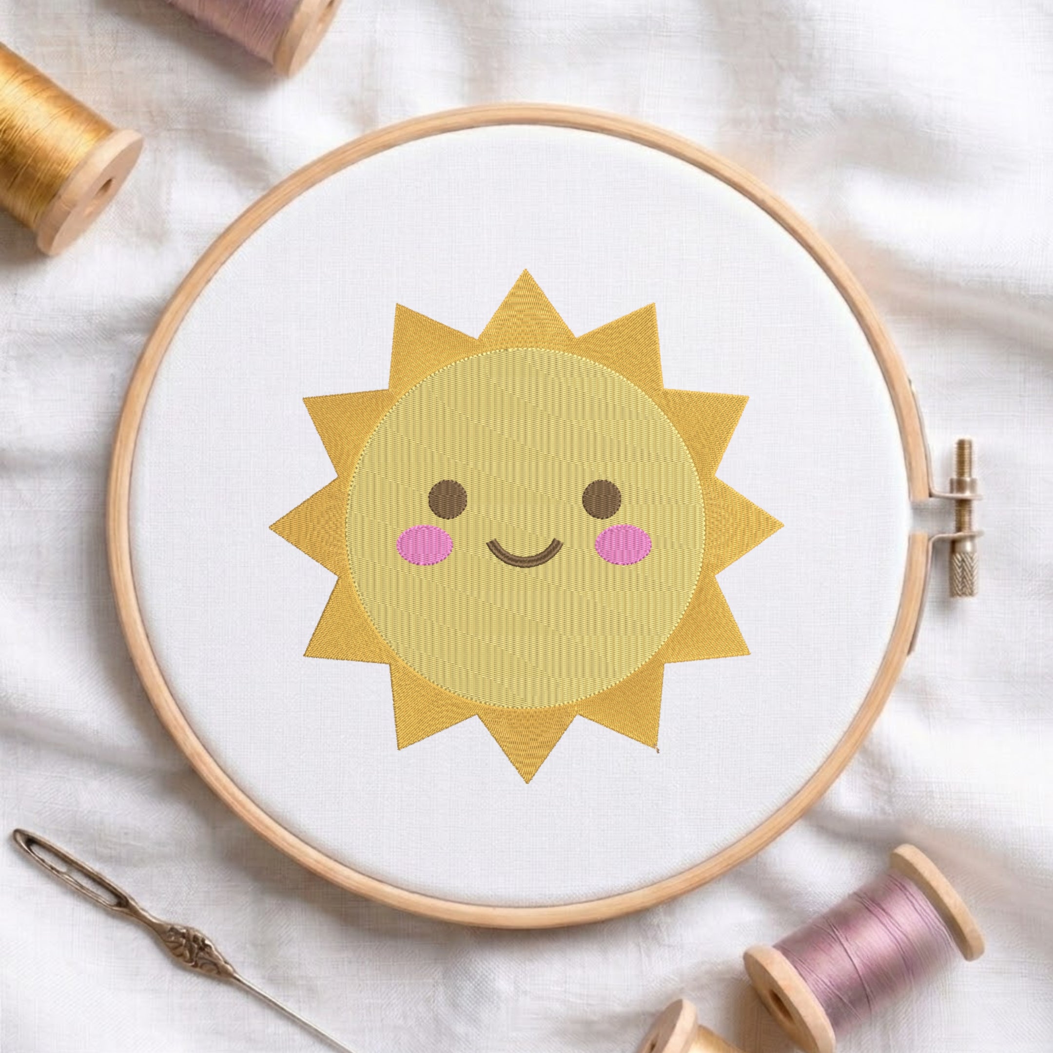 A wooden embroidery hoop holds white fabric with a smiling yellow sun design, surrounded by spools of thread and a needle on a soft white cloth background.