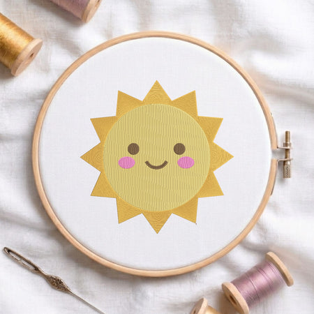 A wooden embroidery hoop holds white fabric with a smiling yellow sun design, surrounded by spools of thread and a needle on a soft white cloth background.