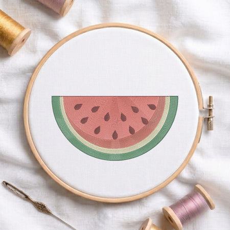 A circular embroidery hoop holding white fabric with a watermelon slice design in pink, green, and beige tones. A needle and thread spools are arranged around it on a white fabric background.
