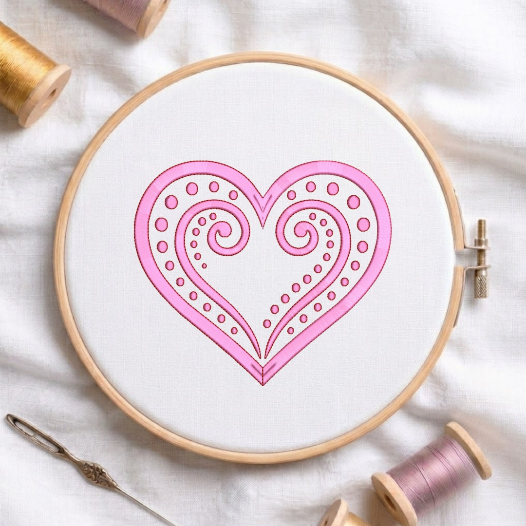 A circular embroidery hoop holding white fabric with a pink heart design outlined in black. A metal needle lies beside the hoop, and spools of thread are arranged around it on a white fabric background.