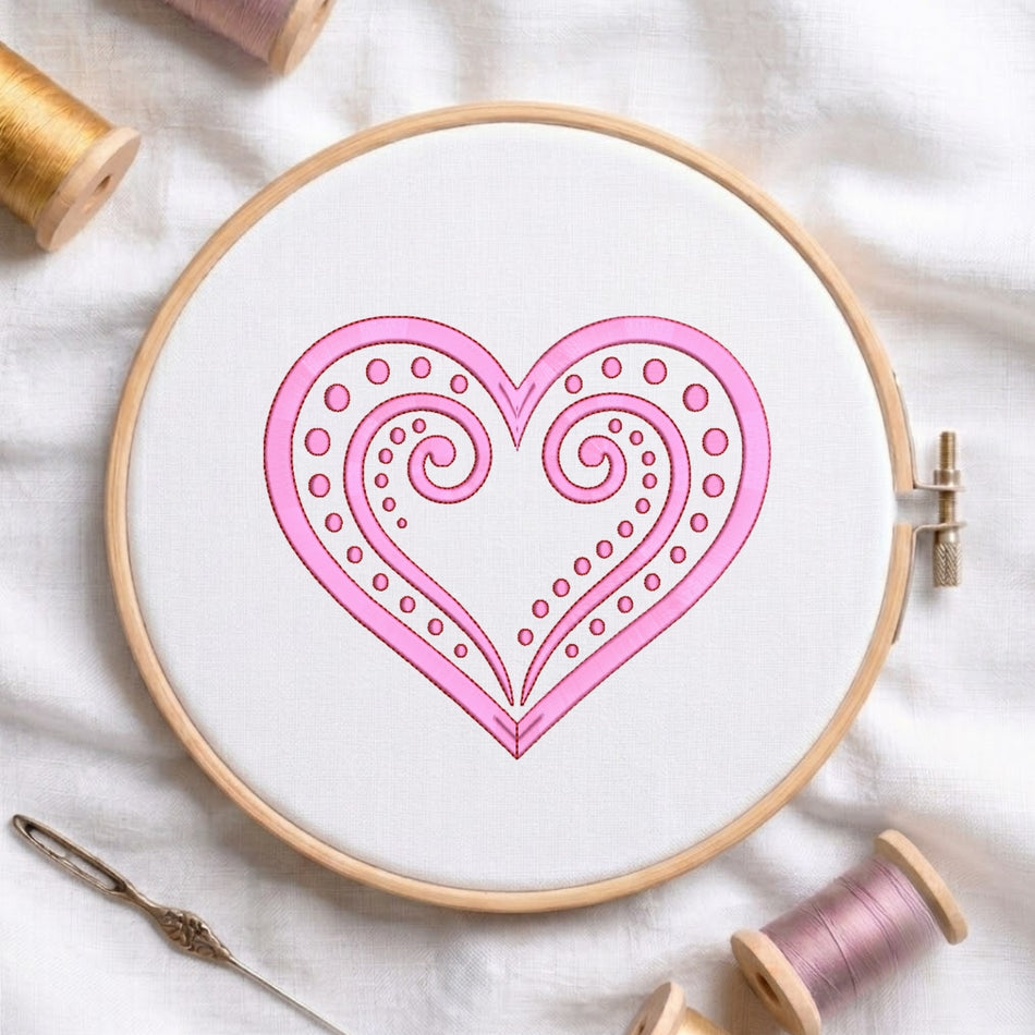 A circular embroidery hoop holding white fabric with a pink heart design outlined in black. A metal needle lies beside the hoop, and spools of thread are arranged around it on a white fabric background.