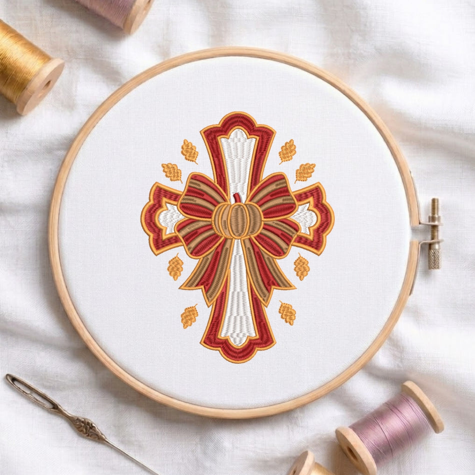 A circular embroidery hoop holding white fabric with a decorative autumn-themed cross design in red, gold, and white. Spools of thread and a needle are arranged around it on a white fabric background.