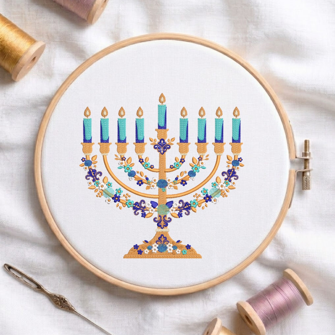 A circular embroidery hoop holding white thread, featuring a colorful embroidered menorah design with blue candles, gold branches, and floral details. The hoop is styled on a white fabric background with spools of thread and a needle nearby.