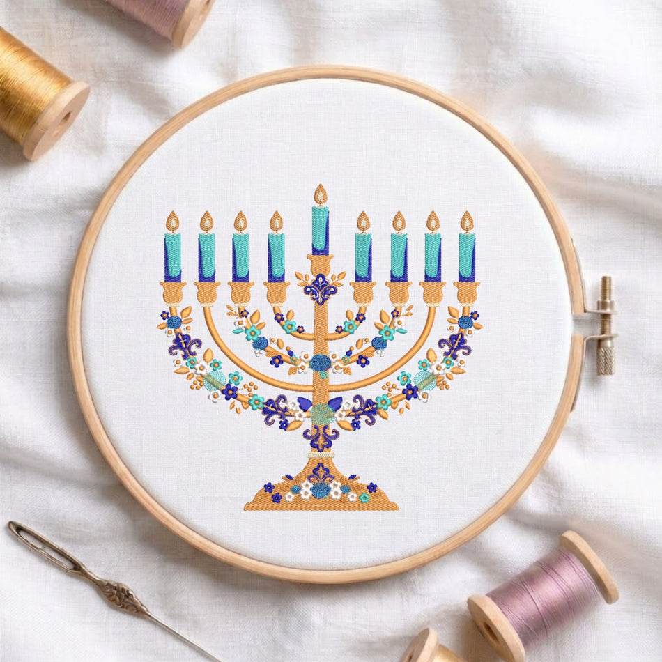 A circular embroidery hoop holding white thread, featuring a colorful embroidered menorah design with blue candles, gold branches, and floral details. The hoop is styled on a white fabric background with spools of thread and a needle nearby.
