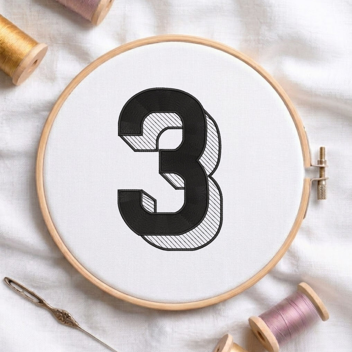 An embroidered 3D block number "3" with striped shading texture, stitched inside a wooden hoop on white fabric with sewing threads.
