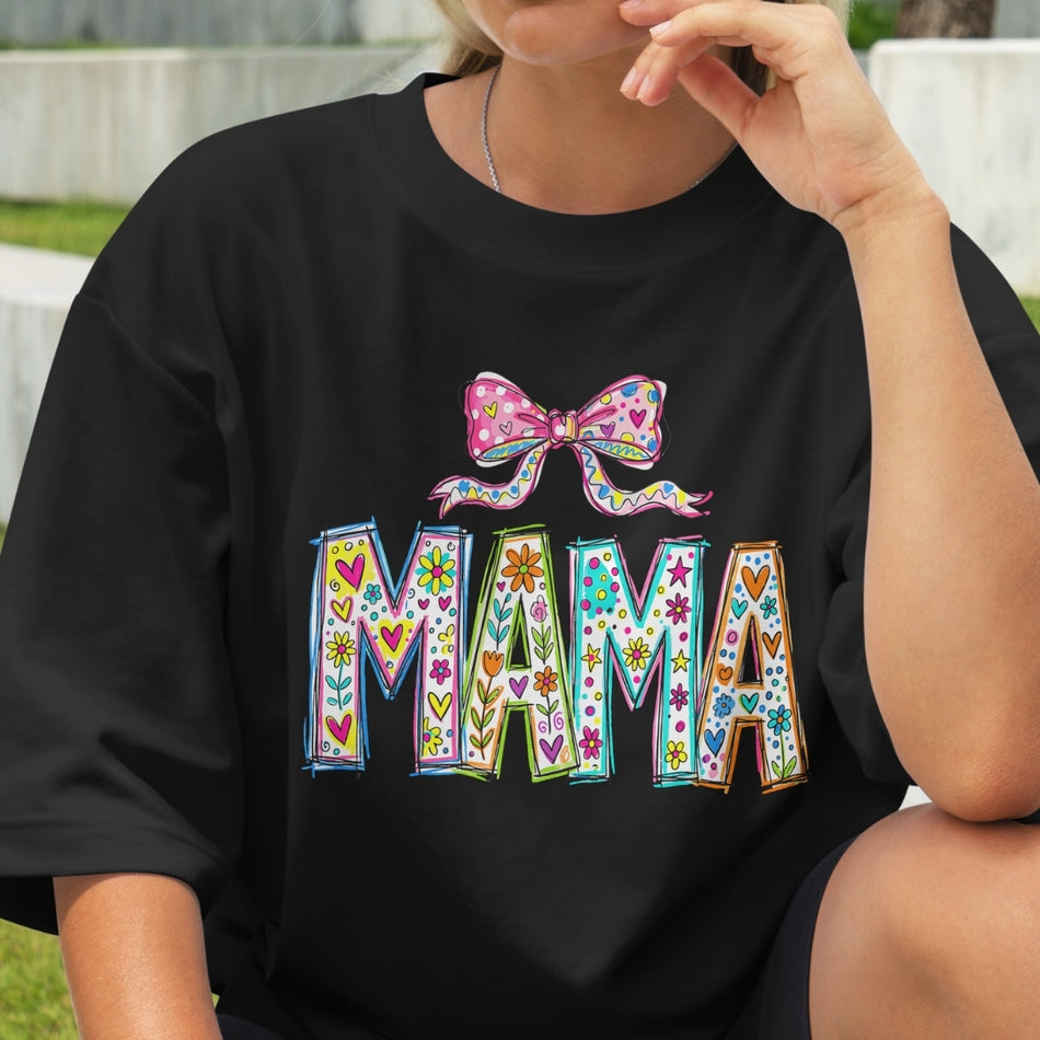 Black t-shirt with colorful 'MAMA' text and bow design worn by a person.