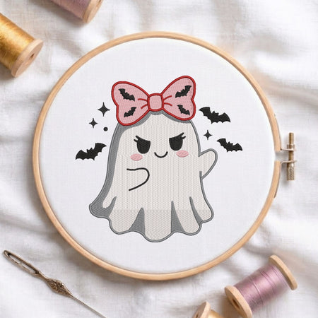 A white fabric stretched in a wooden embroidery hoop, featuring an embroidered design of a ghost with a pink bat-patterned bow, surrounded by flying bats and sparkles.