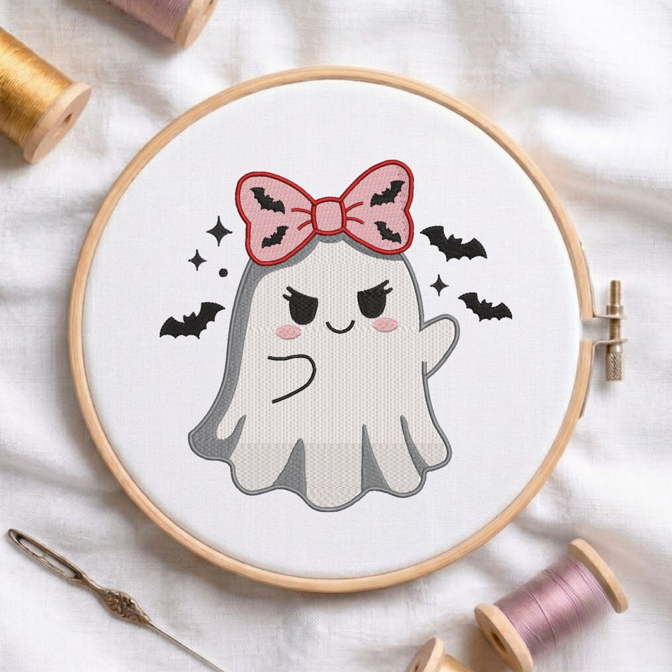 A white fabric stretched in a wooden embroidery hoop, featuring an embroidered design of a ghost with a pink bat-patterned bow, surrounded by flying bats and sparkles.