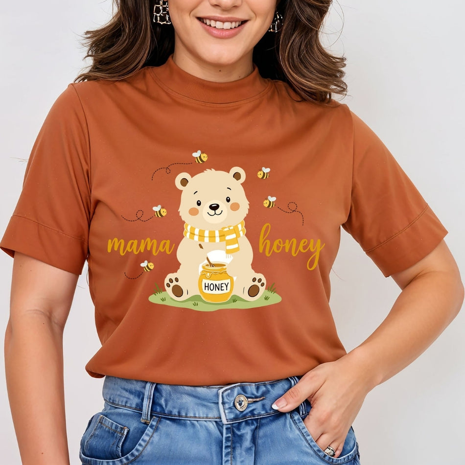 A woman wearing a rust-orange short-sleeve mock-neck t-shirt with a cute cartoon bear graphic holding a jar of honey, surrounded by bees and the text 'mama honey'.