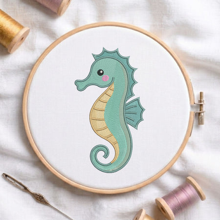 A wooden embroidery hoop holding white fabric with a stitched seahorse design in mint green, pale yellow, and pink. Spools of thread and a needle are arranged around it on a white fabric background.