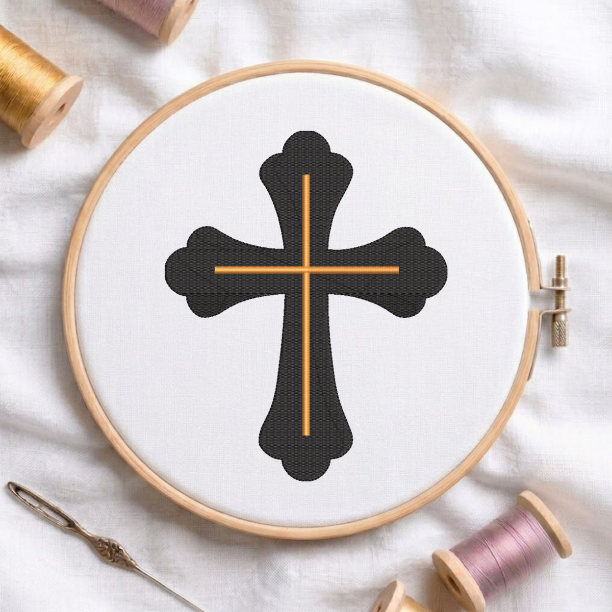 An embroidery hoop on a white cloth displays a finished embroidery design of an ornate black cross with gold intersecting lines, flanked by spools of thread and a needle.