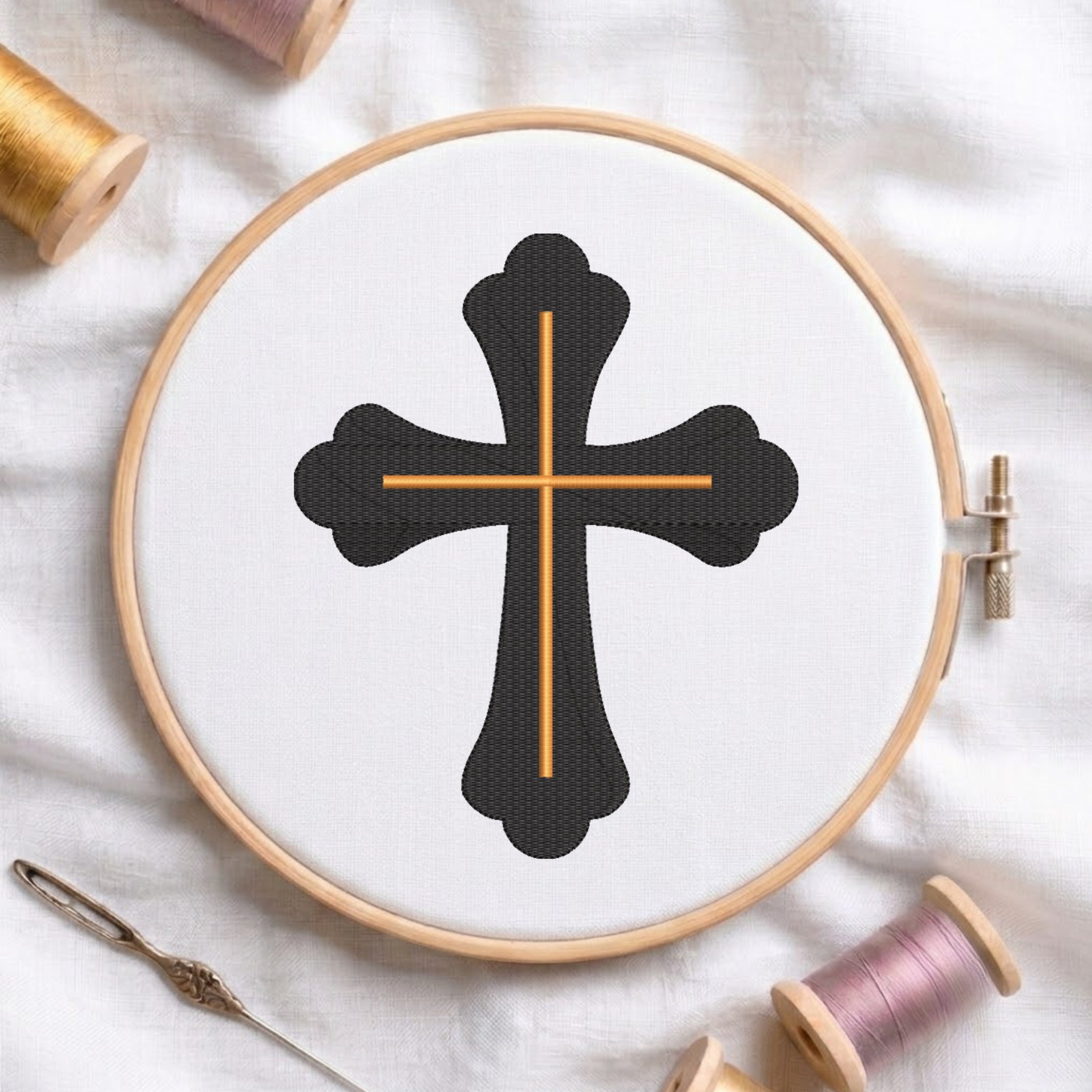 An embroidery hoop on a white cloth displays a finished embroidery design of an ornate black cross with gold intersecting lines, flanked by spools of thread and a needle.