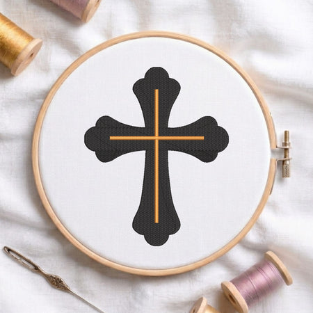 An embroidery hoop on a white cloth displays a finished embroidery design of an ornate black cross with gold intersecting lines, flanked by spools of thread and a needle.