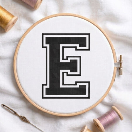 Close-up view of an embroidered varsity-style block letter 'E' in black with a white and black outline, centered in a wooden embroidery hoop surrounded by thread spools and a craft needle.
