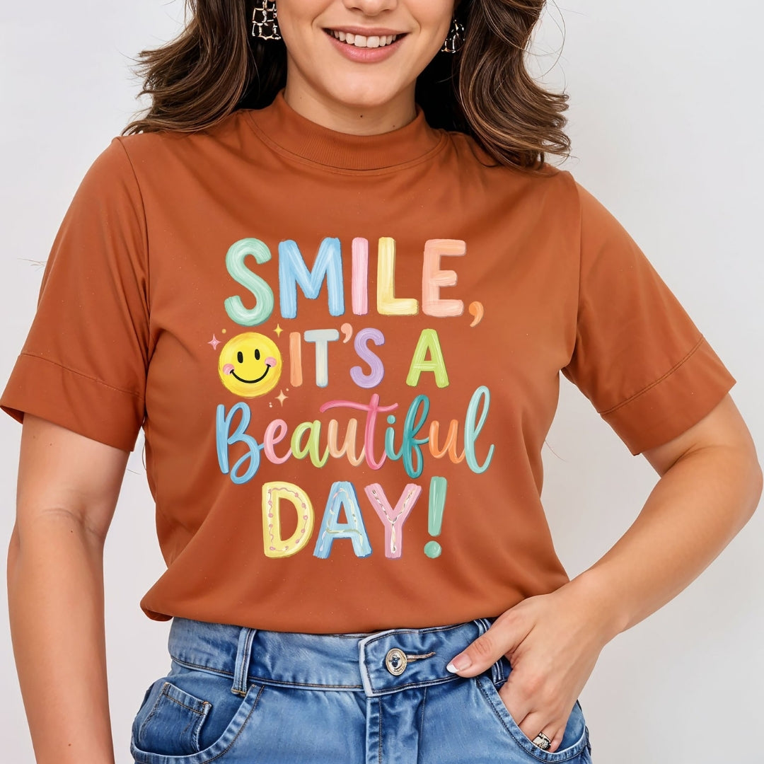 A woman wearing a rust-orange short-sleeve t-shirt with a colorful graphic that reads 'SMILE, IT'S A Beautiful DAY!' in pastel lettering with a yellow smiley face, paired with blue jeans.