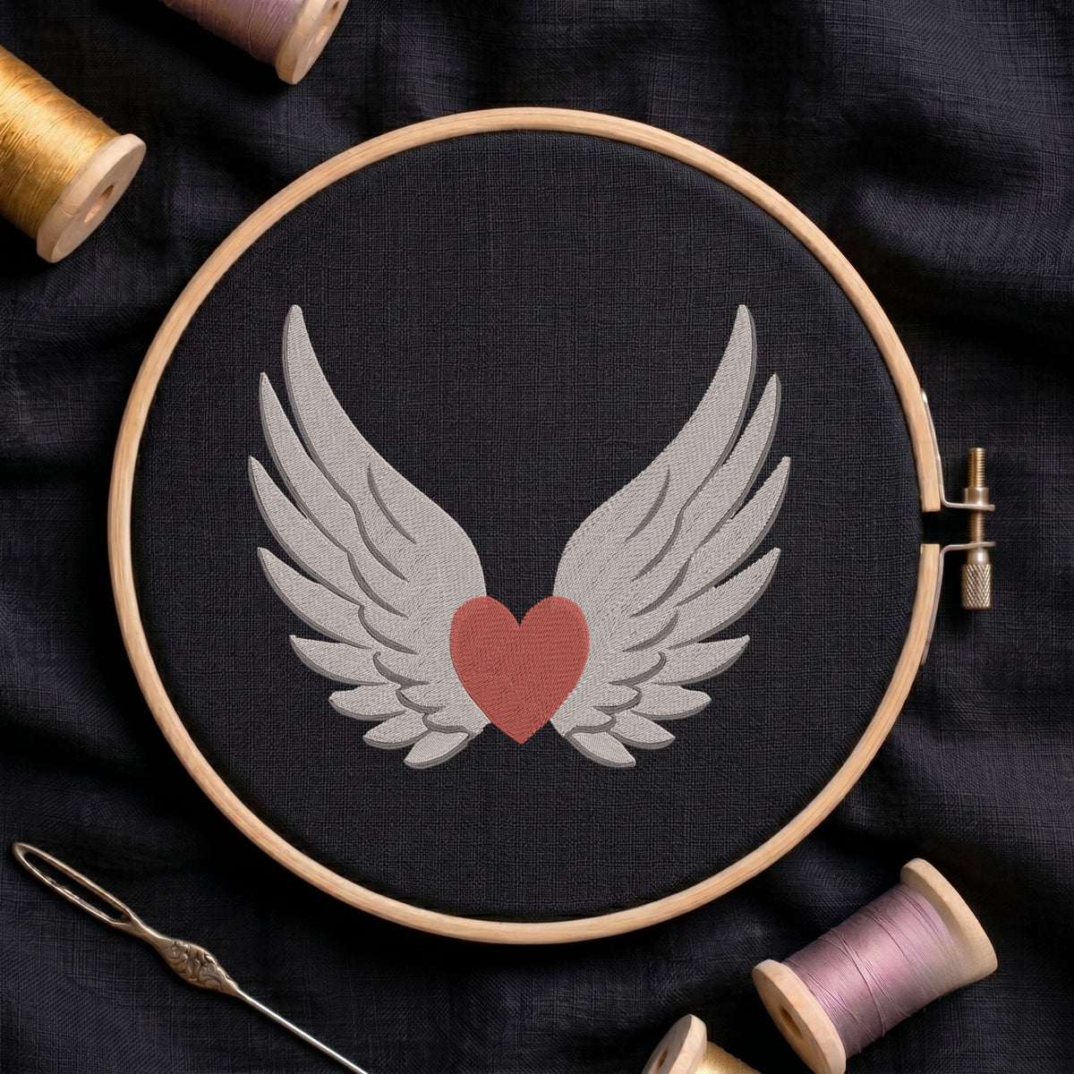 A circular embroidery hoop holding black fabric with a stitched design of white angel wings around a muted red heart. The hoop is photographed from above on dark fabric, with spools of thread and a needle around it.