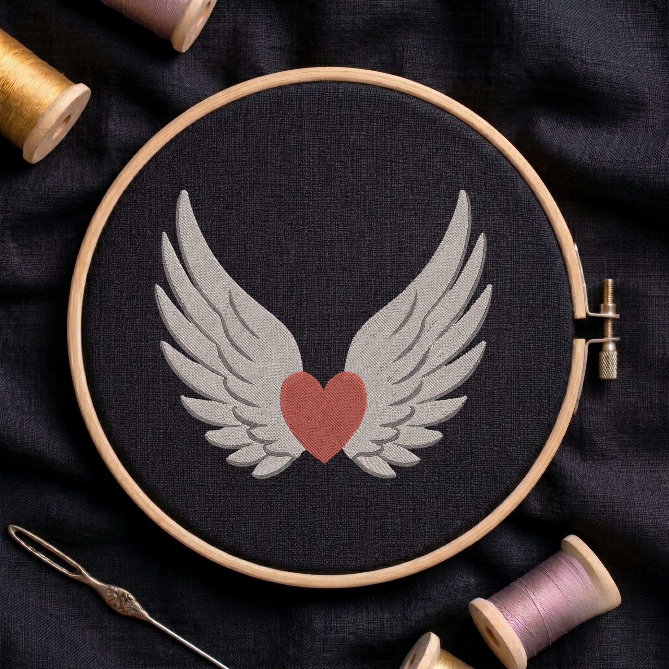 A circular embroidery hoop holding black fabric with a stitched design of white angel wings around a muted red heart. The hoop is photographed from above on dark fabric, with spools of thread and a needle around it.