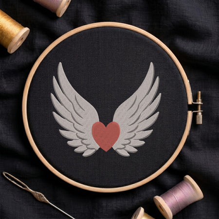 A circular embroidery hoop holding black fabric with a stitched design of white angel wings around a muted red heart. The hoop is photographed from above on dark fabric, with spools of thread and a needle around it.