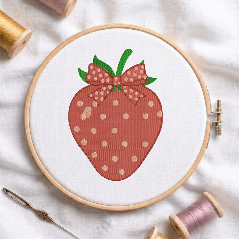 A strawberry embroidery design held in a wooden embroidery hoop, surrounded by spools of thread and a needle on a white fabric background.