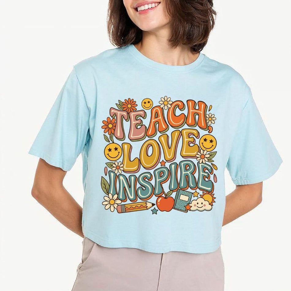 Smiling person wearing a light blue cropped t-shirt featuring the retro "TEACH LOVE INSPIRE" graphic and bold, groovy typography.