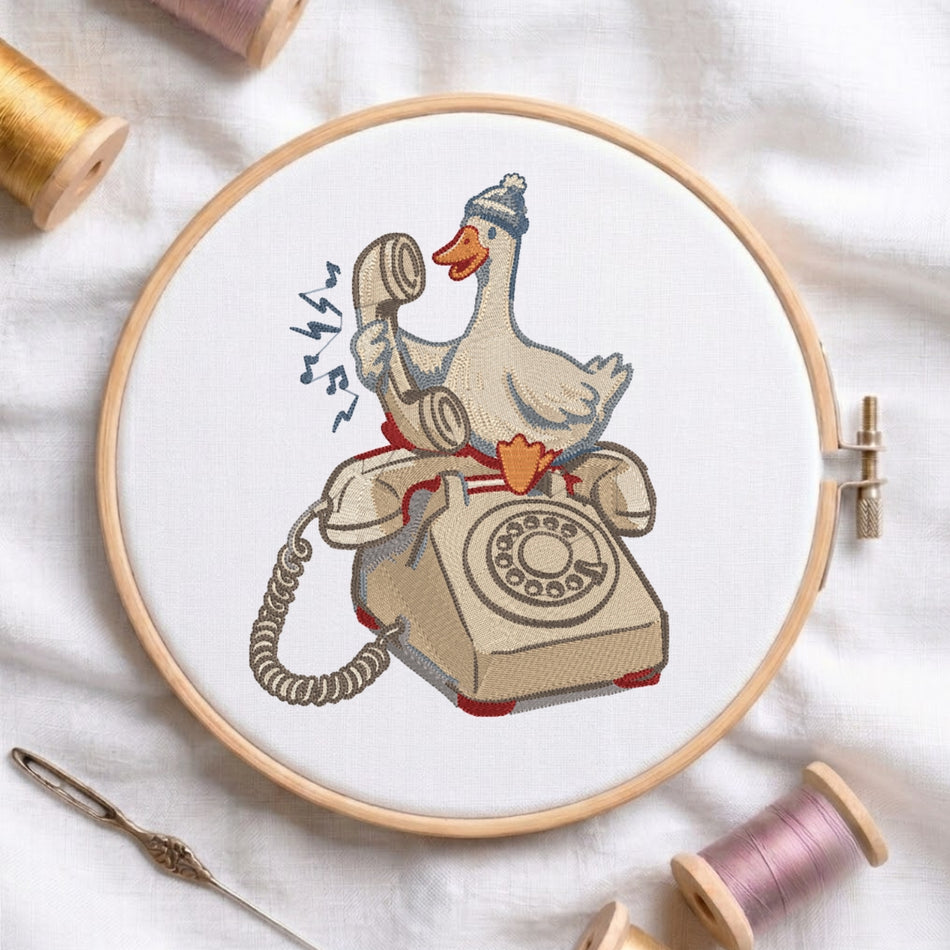 A circular embroidery hoop on white fabric displays a stitched design of a duck wearing a beanie and sitting on a rotary telephone, surrounded by spools of thread and a needle.