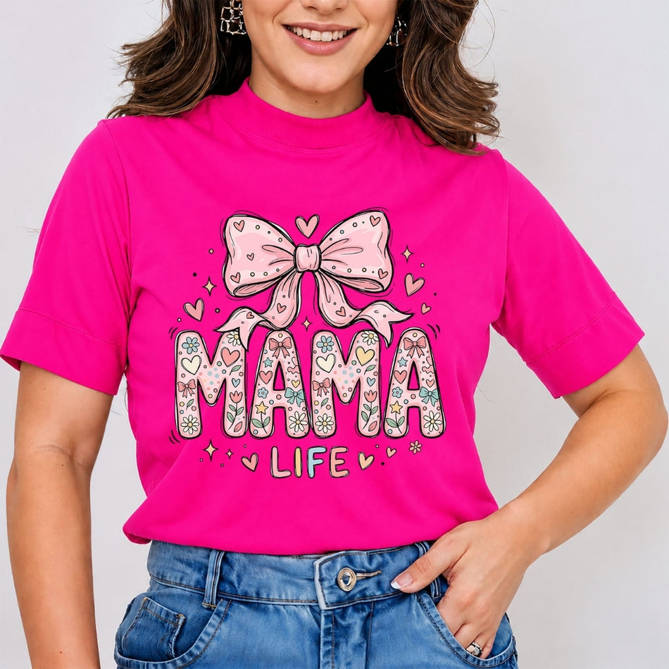 Woman wearing a vibrant hot pink mock neck t-shirt featuring a cute coquette style "MAMA LIFE" graphic text filled with floral patterns and topped with a large pink bow, tucked into blue jeans.