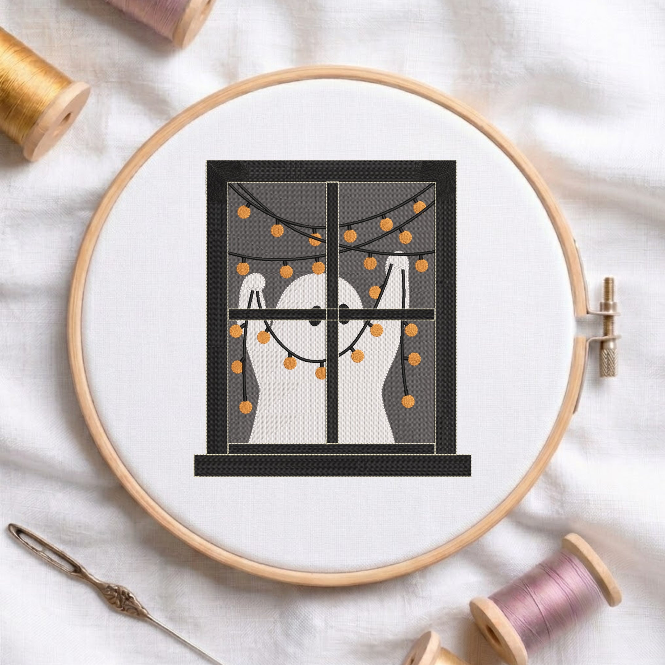 A circular embroidery hoop on white fabric, featuring a stitched design of two white ghosts behind a window with orange string lights. Surrounding props include spools of thread and a needle.